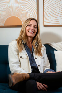 Kelli Russell smiling warmly on a navy couch with sunburst and abstract artwork behind her